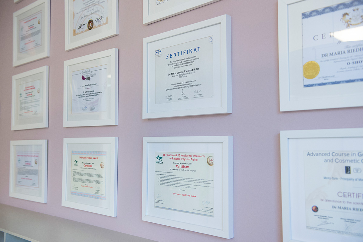 Eine Gruppe gerahmter Zertifikate von Dr. Maria Riedhart-Huter an einer Wand.