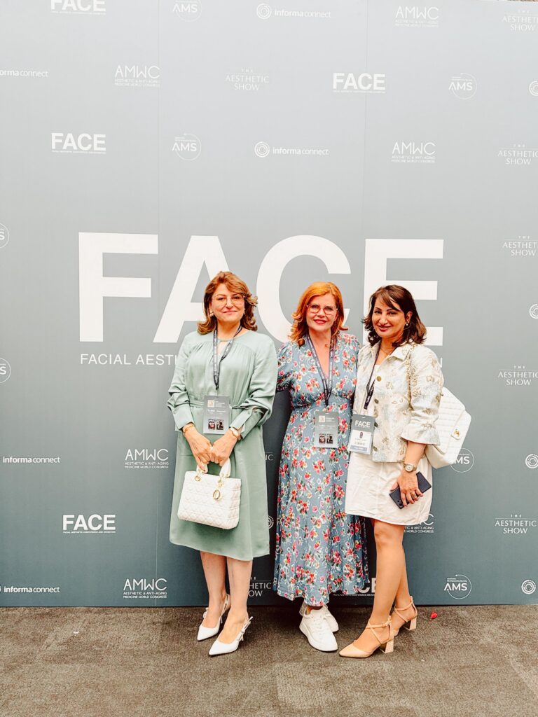 Drei Ärztinnen oder Fachfrauen der ästhetischen Medizin auf der FACE Facial Aesthetic Conference and Exhibition 2025 in London – Gruppenfoto vor der offiziellen Event-Wall. Teilnehmerinnen tragen elegante Outfits mit Namensschildern und Besucherausweisen. Internationale Fachmesse für ästhetische Behandlungen, Anti-Aging und kosmetische Dermatologie, organisiert von Informa Connect und AMWC.